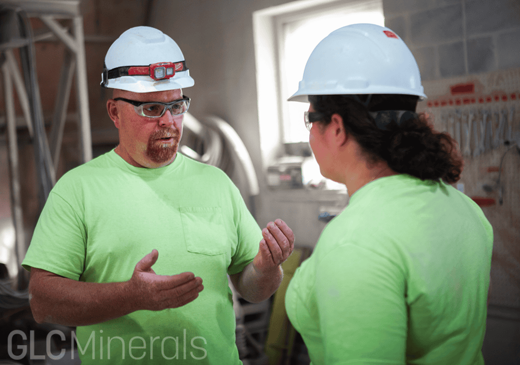 GLC Minerals employees wearing hard hats and safety glasses discuss workplace safety inside the plant.